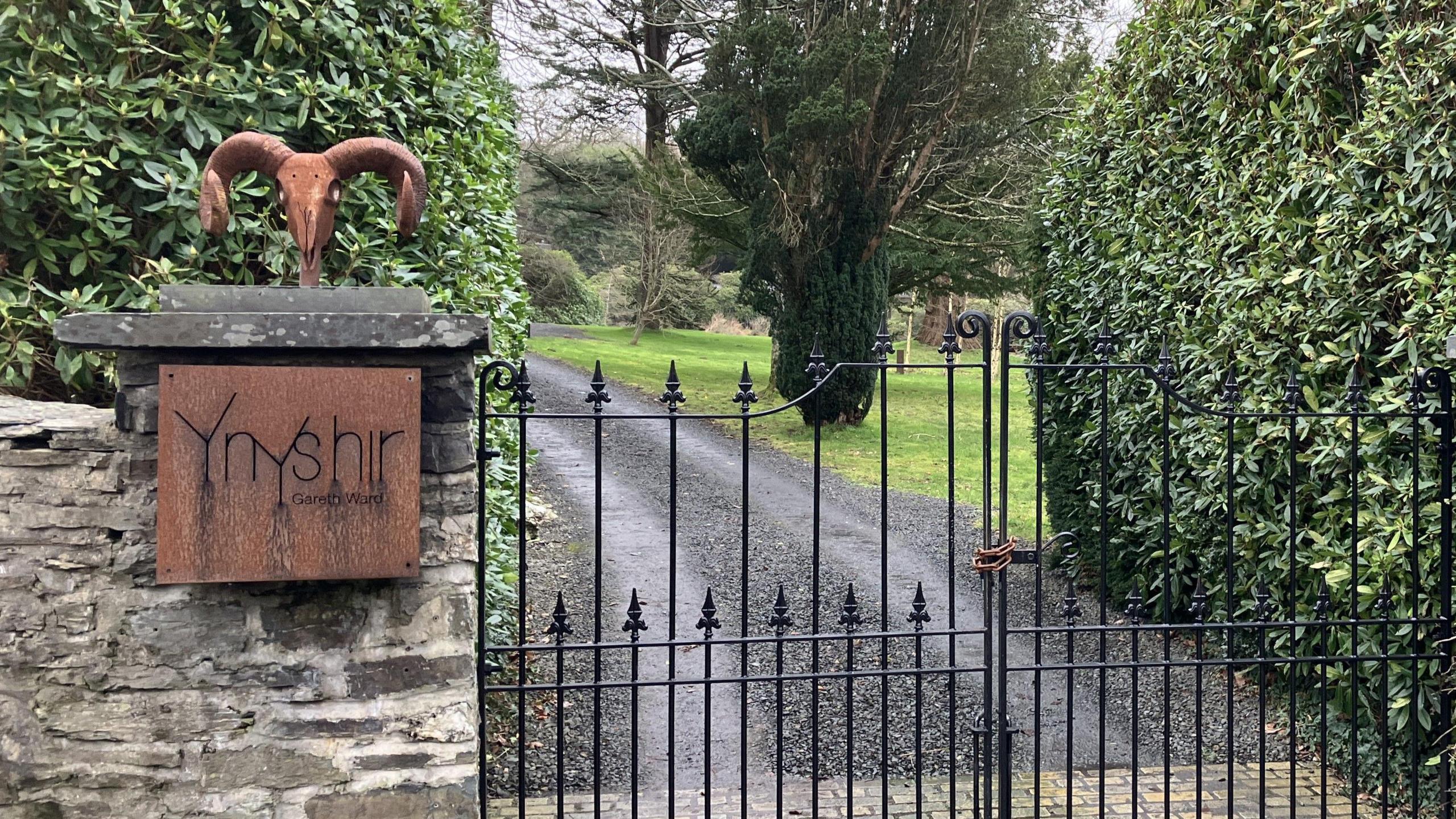 The gated entrance to Ynshir restaurant. A black cast iron gate leads up a drive. On the wall is a bronze coloured sign with the restaurant's name on it.