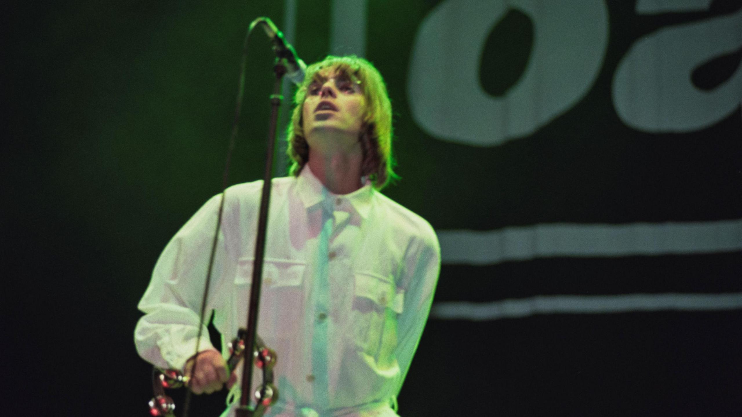 A blurry image shows Liam Gallagher on stage at Knebworth, singing into a microphone on a tall stand and holding a tambourine