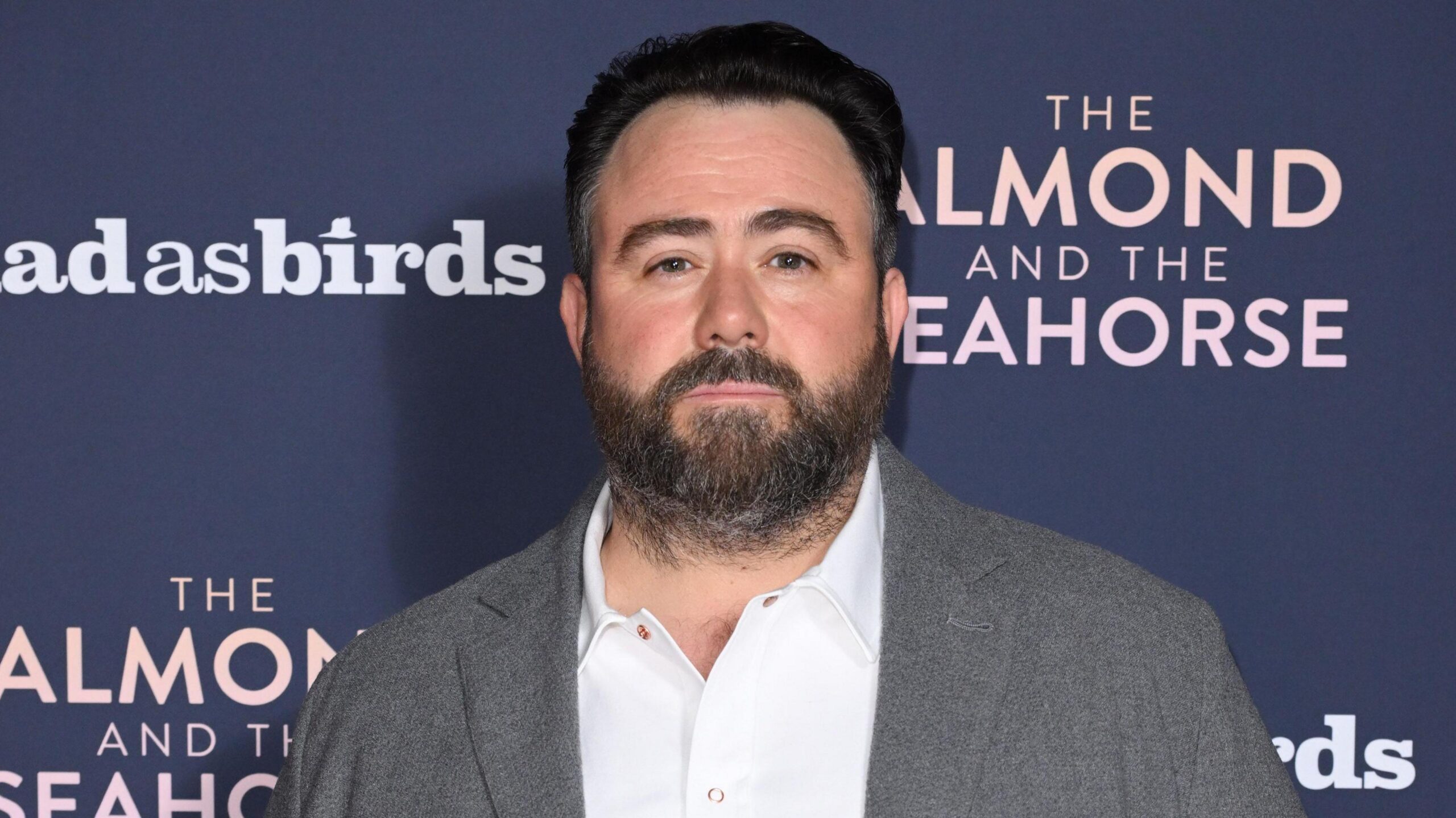 Celyn Jones wearing a grey blazer and a white shirt. He is standing in front of a dark blue screen. He has short dark hair and a short beard.