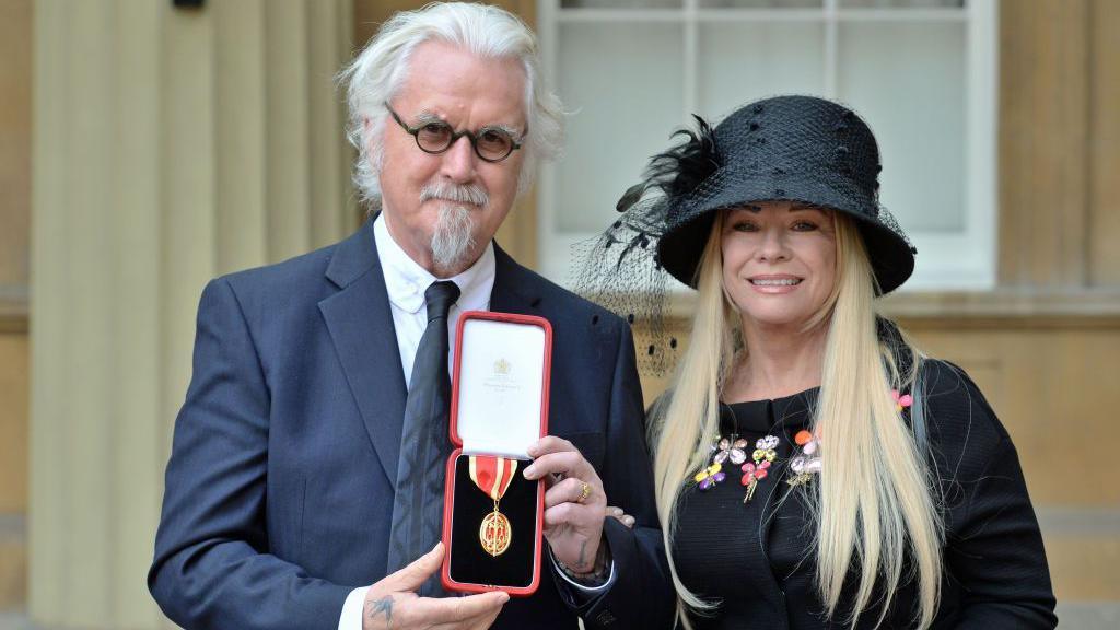Billy Connolly showing his medal to the camera, after being knighted. He has white hair, is wearing a suit and glasses, while smiling. He is standing outside Buckingham Palace with his wife Pamela Stephenson, who has long blonde hair, is wearing a black dress and hat, and is also smiling for the camera.