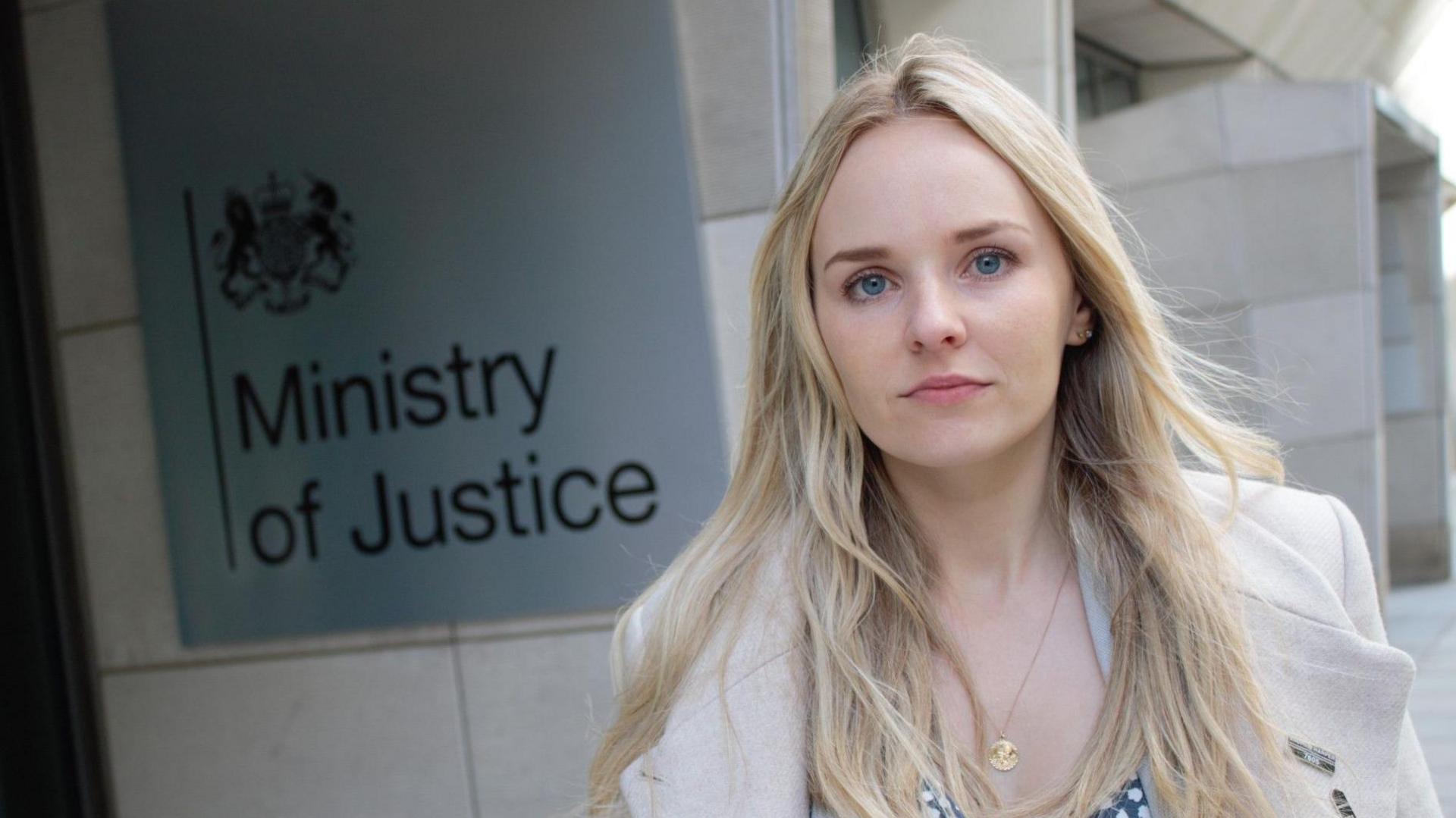 Harper has long blonde hair and blue eyes. She wears a white coat and stands next to the sign for the Ministry of Justice.