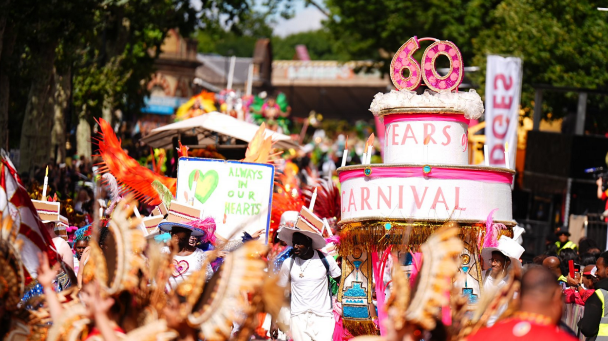 A parade of people walk down the street, with one group holding a large fake birthday cake that reads "60 years of Carnival"