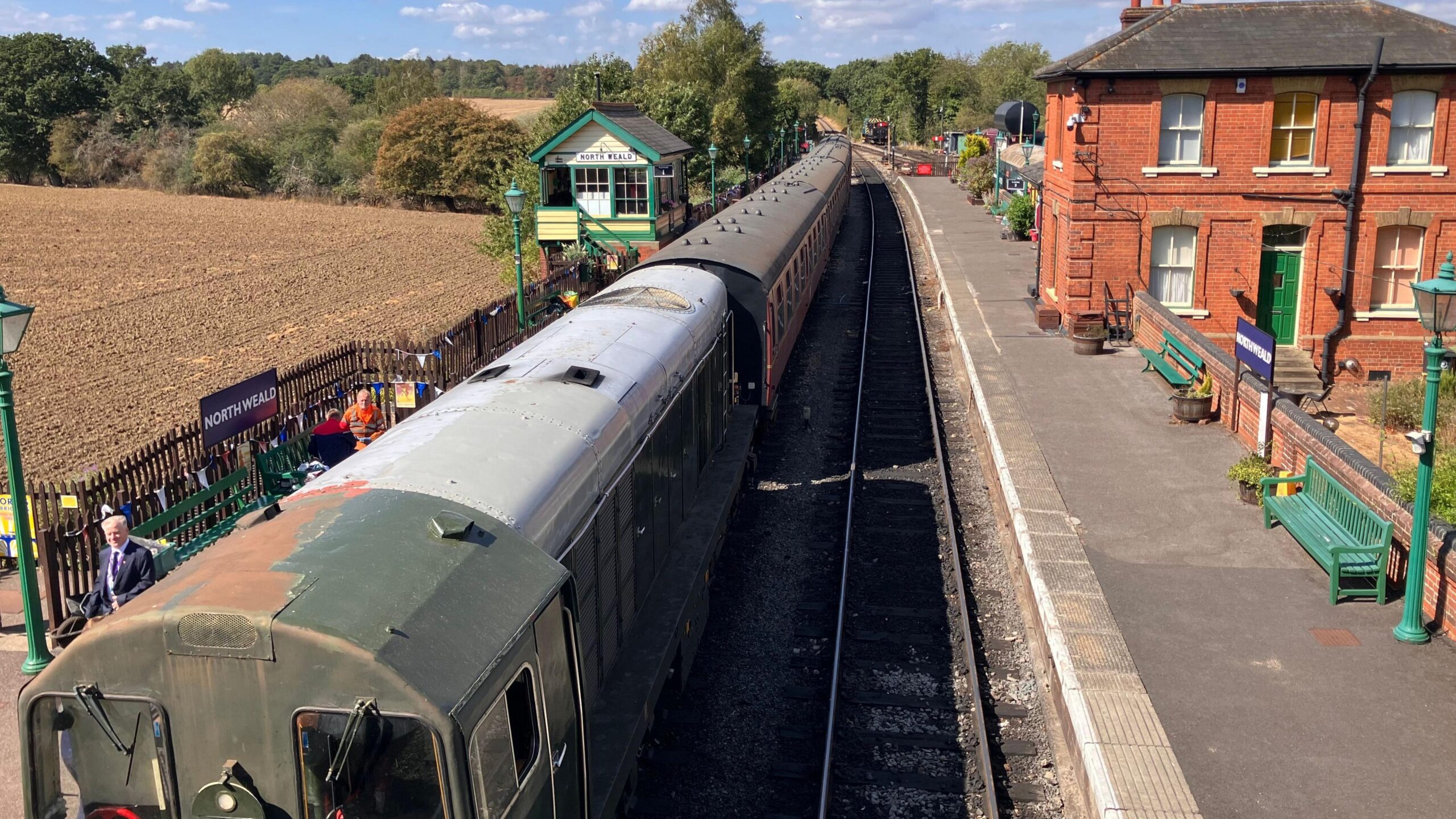 A bird's eye view of the Mark 1 train stationed in North Weald on a sunny day. There are green wooden benches and lamp-posts.