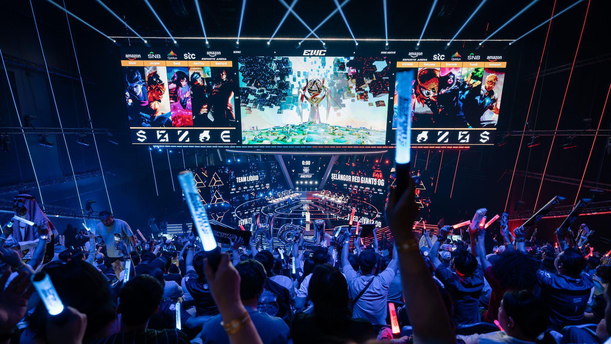 The stage at the Esports World Cup has three overhead screens and a large floor with two teams on each side. A seated crowd is seen holding up their arms in support