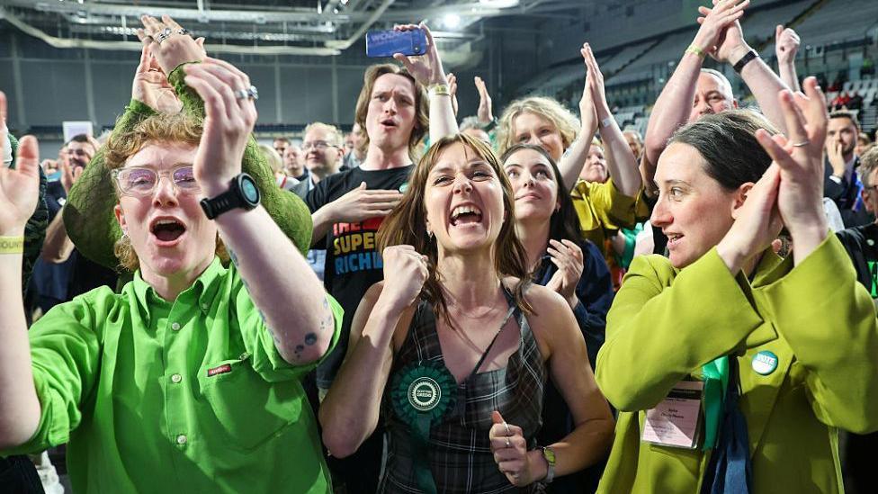 A group of young people dressed in green are all smiling and waving there arms in the air as they celebrate
