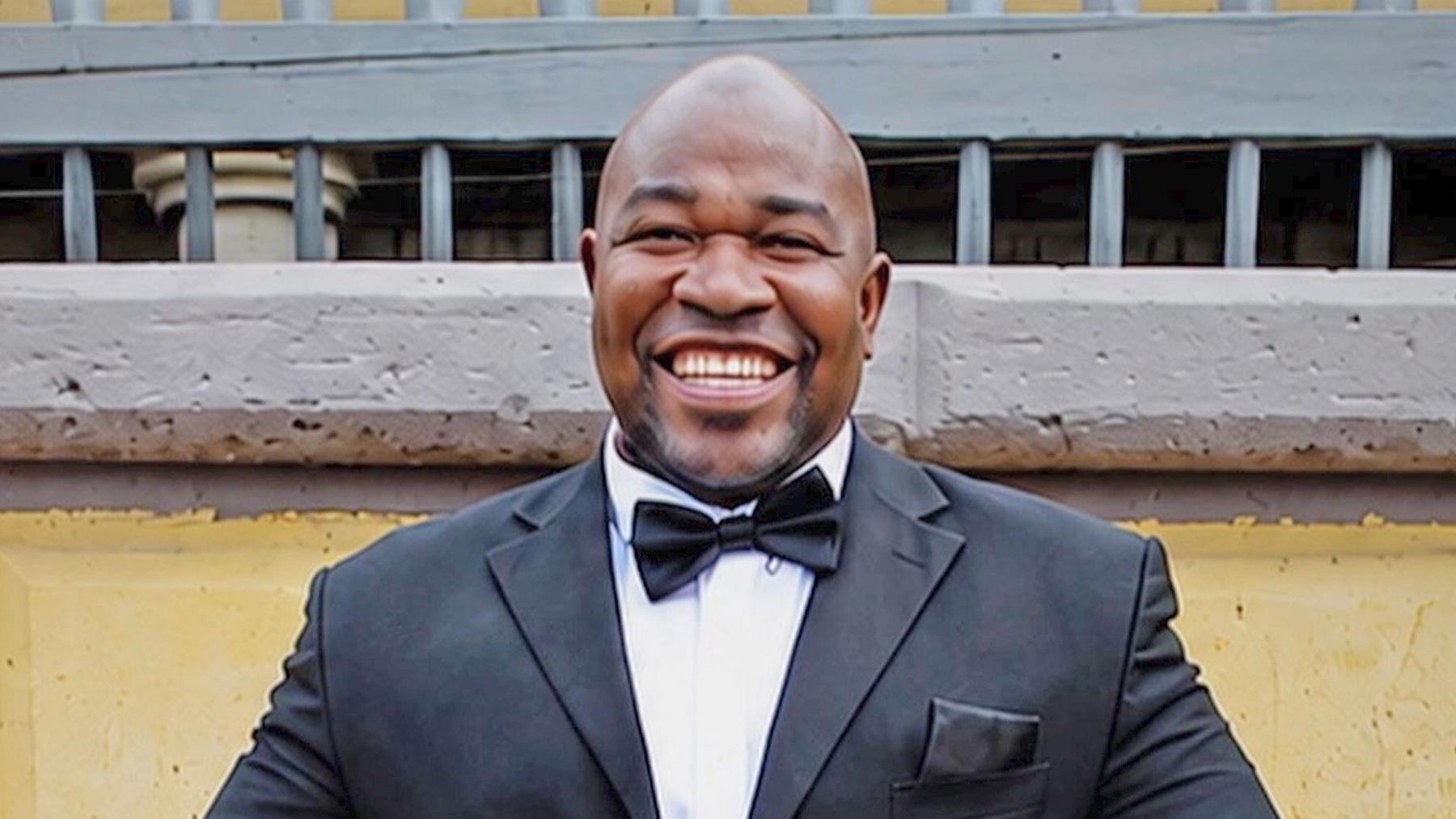 A man smiles broadly at the camera, wearing a black tuxedo and white shirt against a yellow and grey wall
