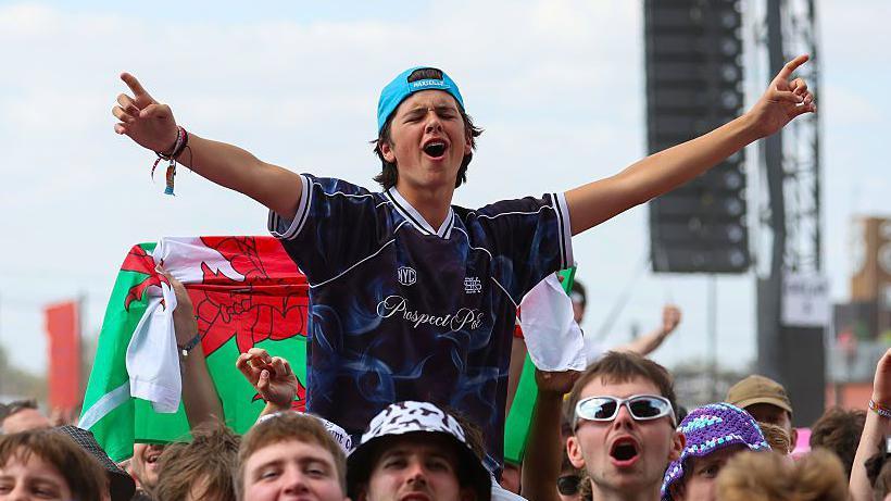 A boy, wearing a blue cap and blue T-shirt, being lofted above on a person's shoulders, who is spreading his arms out wide as he sings to a song at the festival.