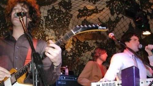 The Killers performing in a dark club in 2003. A man wearing a white shirt sings into a microphone, while a guitarist with long curly hair sings along and a keyboardist plays in front of a wall decorated with a psychedelic design.