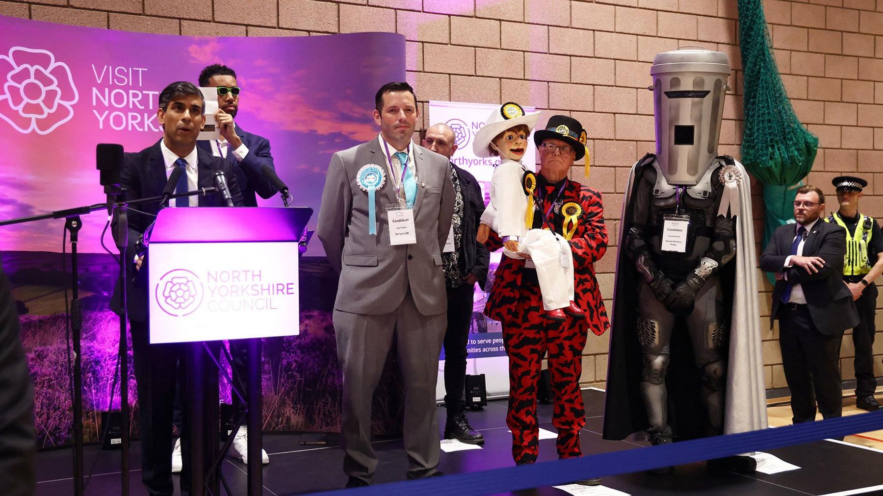 Rishi Sunak is standing on stage in front of a bright purple backdrop with the slogan visit North Yorkshire. He is on the left of the photograph, giving a speech at a podium, with other candidates lined up to his right. Count Binface is standing on the right of the stage in his costume.
