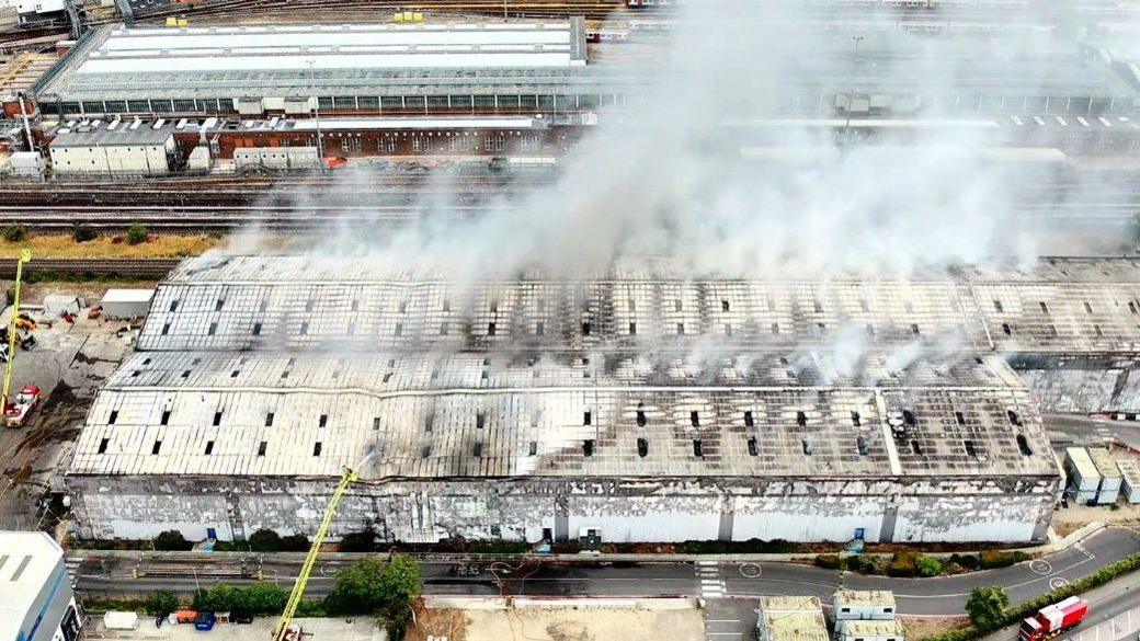 An aerial view of a large industrial warehouse unit with smoke billowing from its roof, while a London Fire Brigade turntable ladder directs water onto the building from below.