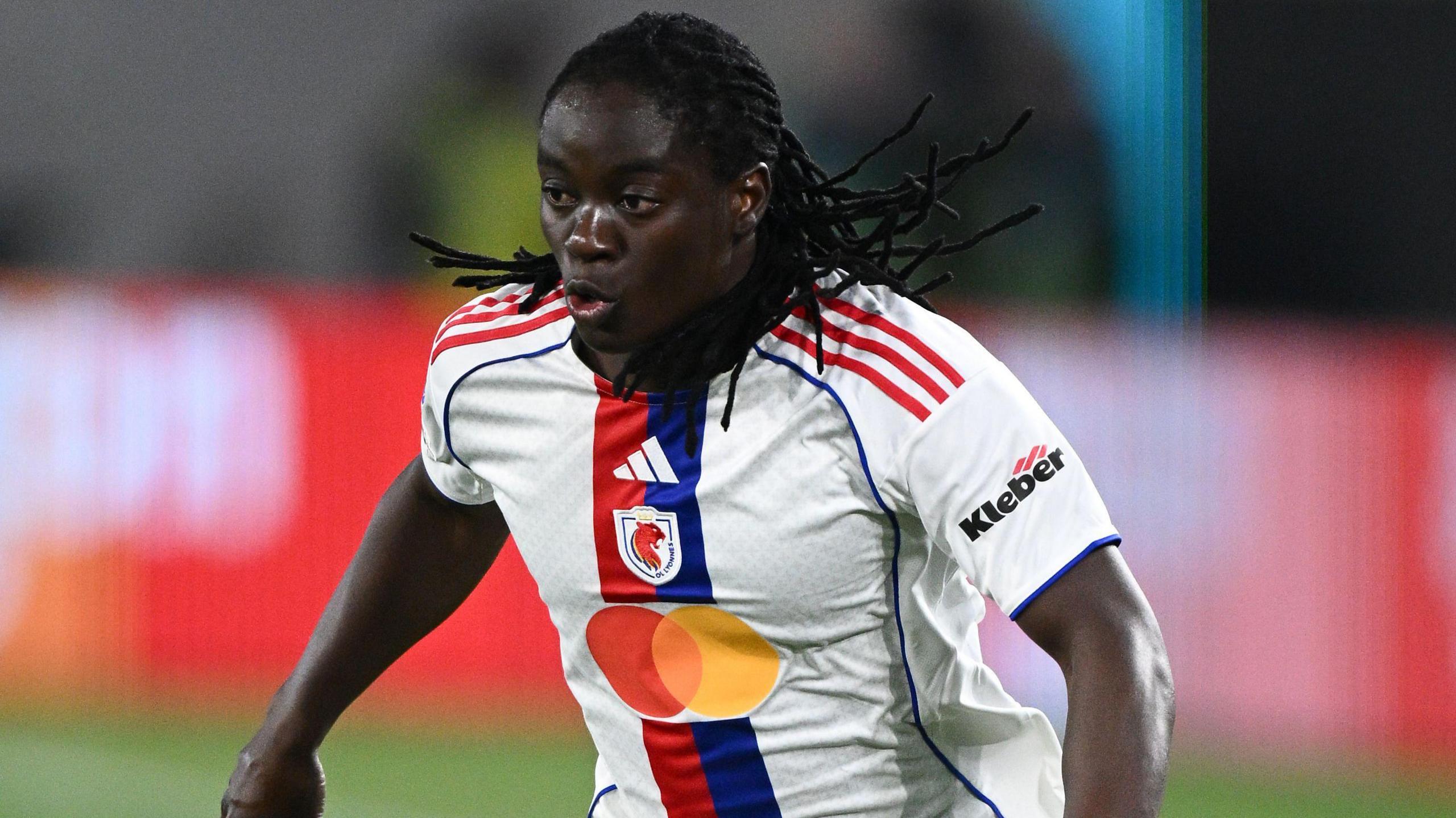 Tabitha Chawinga visible from waist up while in action wearing a white Lyon shirt with red and blue detail running down its centre. She puffs out her cheeks as she runs during a football match, with her many braids flapping in all directions behind and to the side of her head