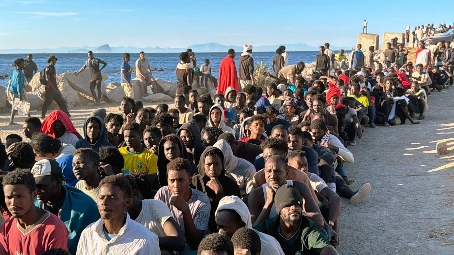 Migrants sitting on the ground in rows at the beach with the sea behind them in Ceuta waiting for aid