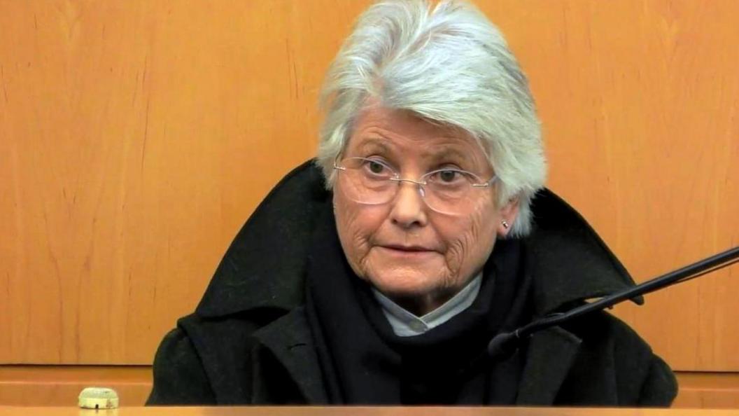 A 77-year-old woman with white hair and glasses, wearing a black coat, sits in the witness box at the High Court in Glasgow