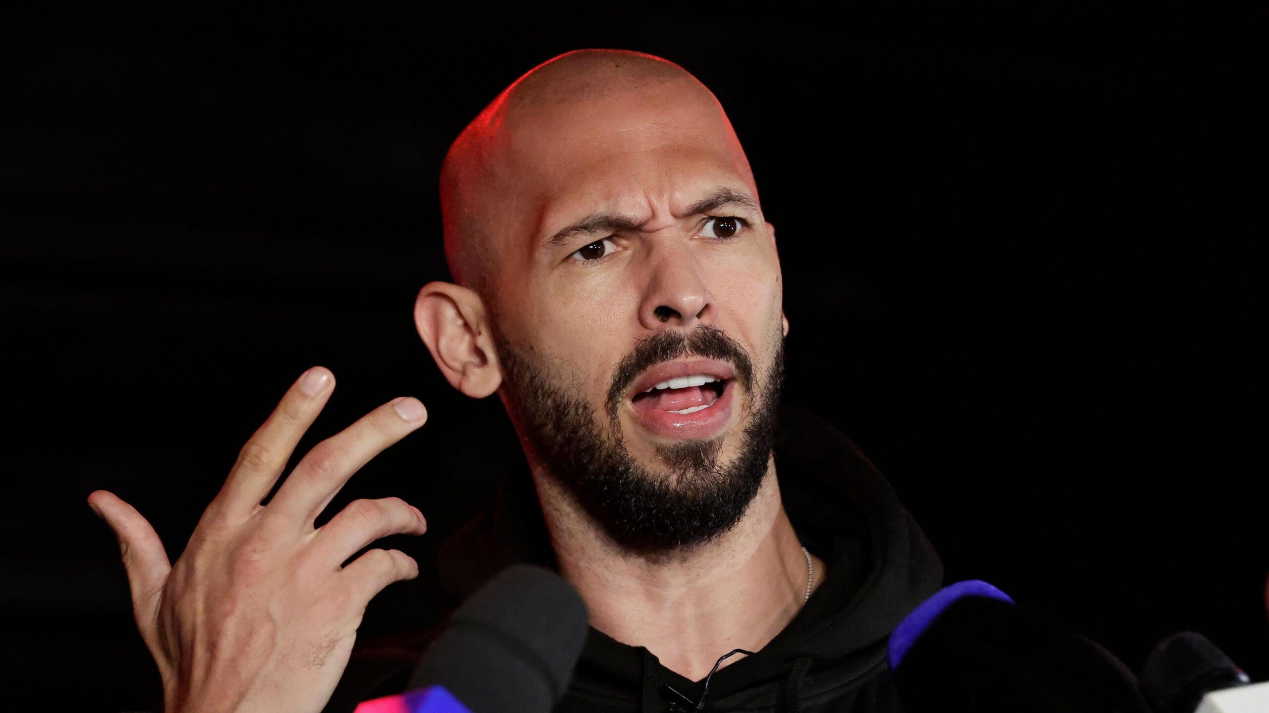 Andrew Tate speaking to the media in March 2025. He gestures with his hand and speaks while several microphones are pointed towards him.