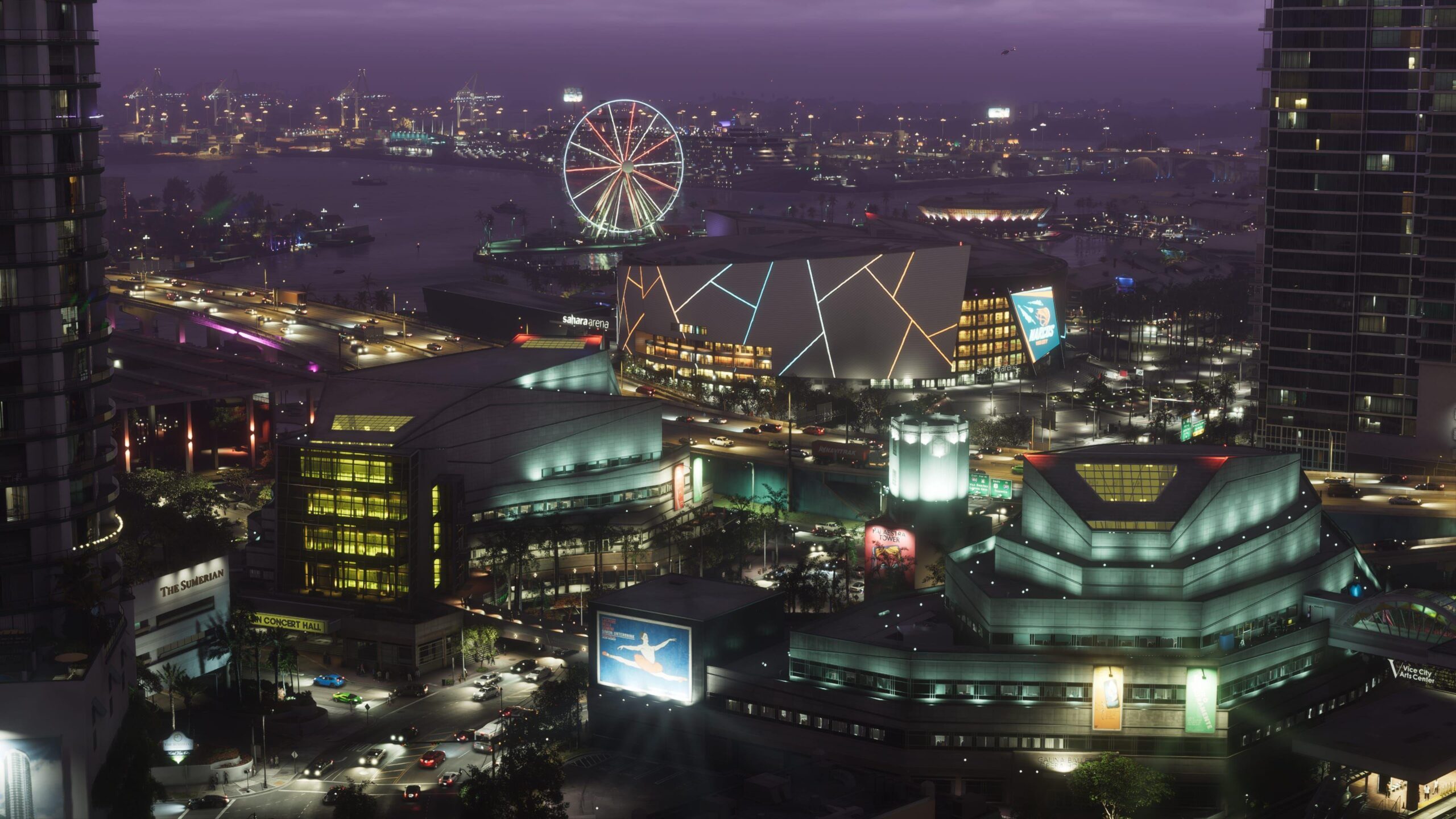 An aerial screenshot shows downtown Vice City at night. Blocks of apartments, a stadium with a large animated billboard and an illuminated Ferris wheel create a glowing tapestry of colours. In the distance, on the other side of a port, the lights of more buildings dot the skyline.