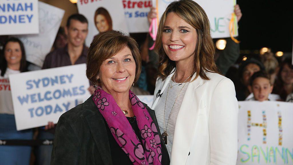 Savannah, all in white, and Nancy Guthrie in a black suit jacket and pink patterned scarf, smile as they stand shoulder to shoulder. People behind them hold up signs.