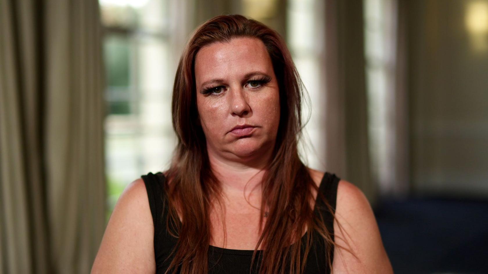 A woman wearing a black tank top and long reddish brown hair sits and stares at the camera.