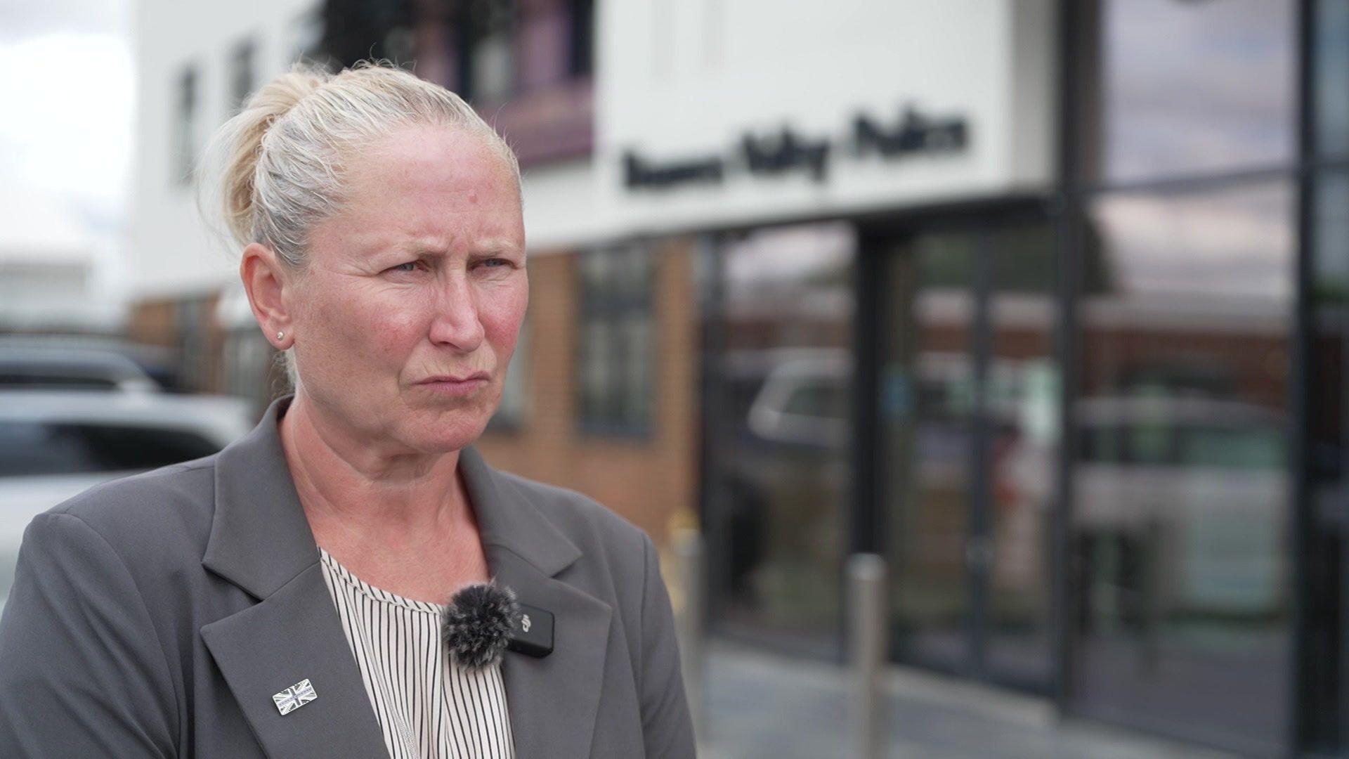 Aileen O'Connor has blonde hair in a bun and is wearing a grey blazer over a black and white shirt. Behind her is Thames Valley Police headquarters.