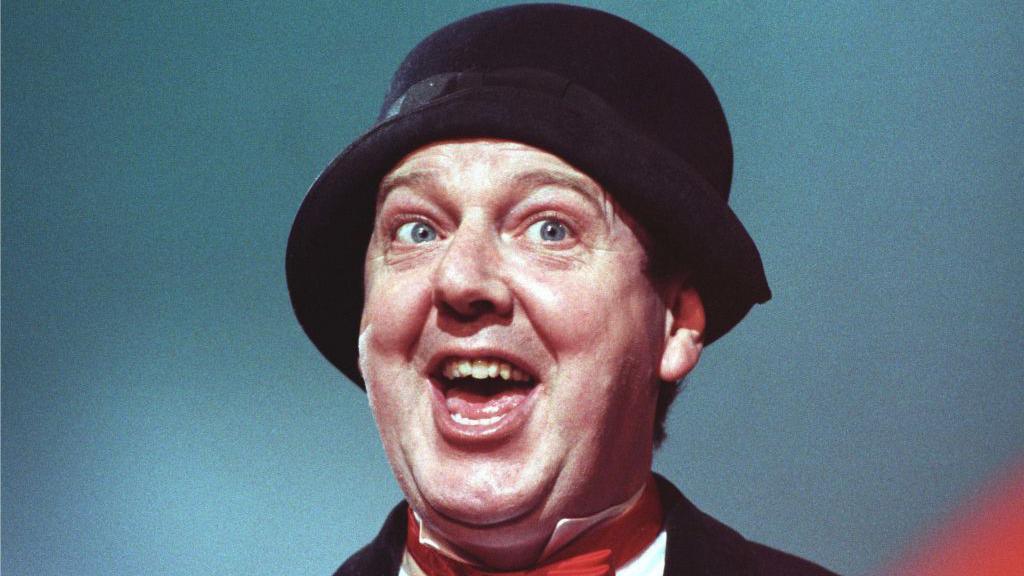 A man wears a black hat that covers the top of his ears. The photo is taken from slightly below him. He looks of to the right, mouth open, blue eyes. He wears a black suit, white shirt and red bow tie.