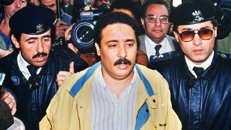Fhimah (centre) being escorted by security officers and surrounded by journalists before a hearing in 1992 in connection with the Lockerbie bombing. He has short brown hair and a moustache and is wearing a yellow jacket and a white and blue striped short.