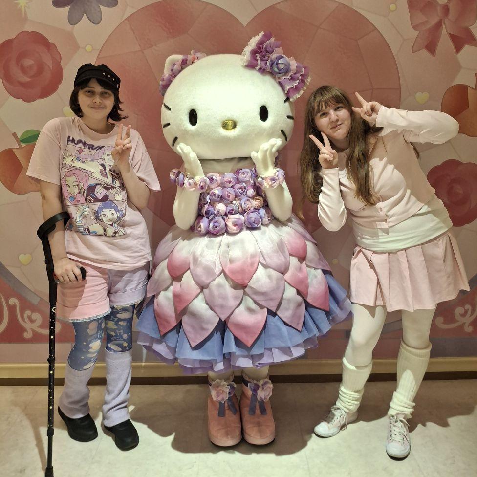 Two people in pastel colours stand next to a person in a Hello Kitty costume