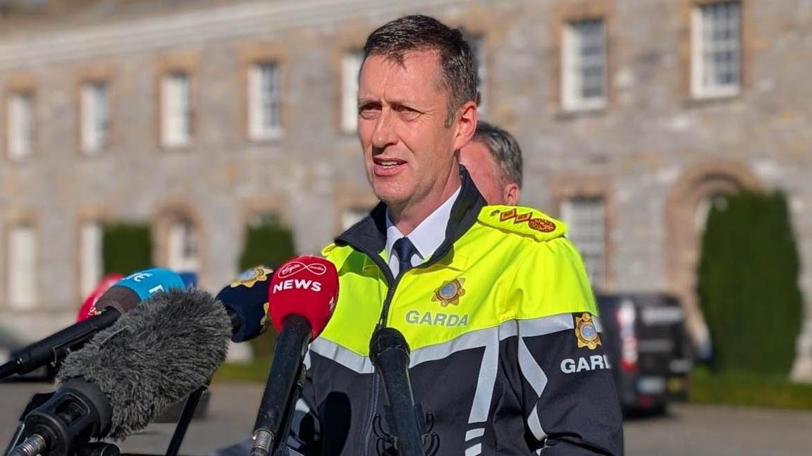 Garda Commissioner Justin Kelly syanding in front of microphones. He has short dark hair, wearing a white shirt, navy tie and high viz garda jacket.