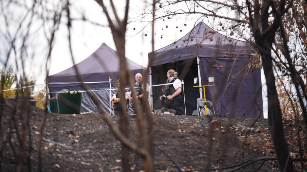 Two temporary tents and several people inside a taped-off area on a hillside with dark ground and sparse vegetation in the foreground.