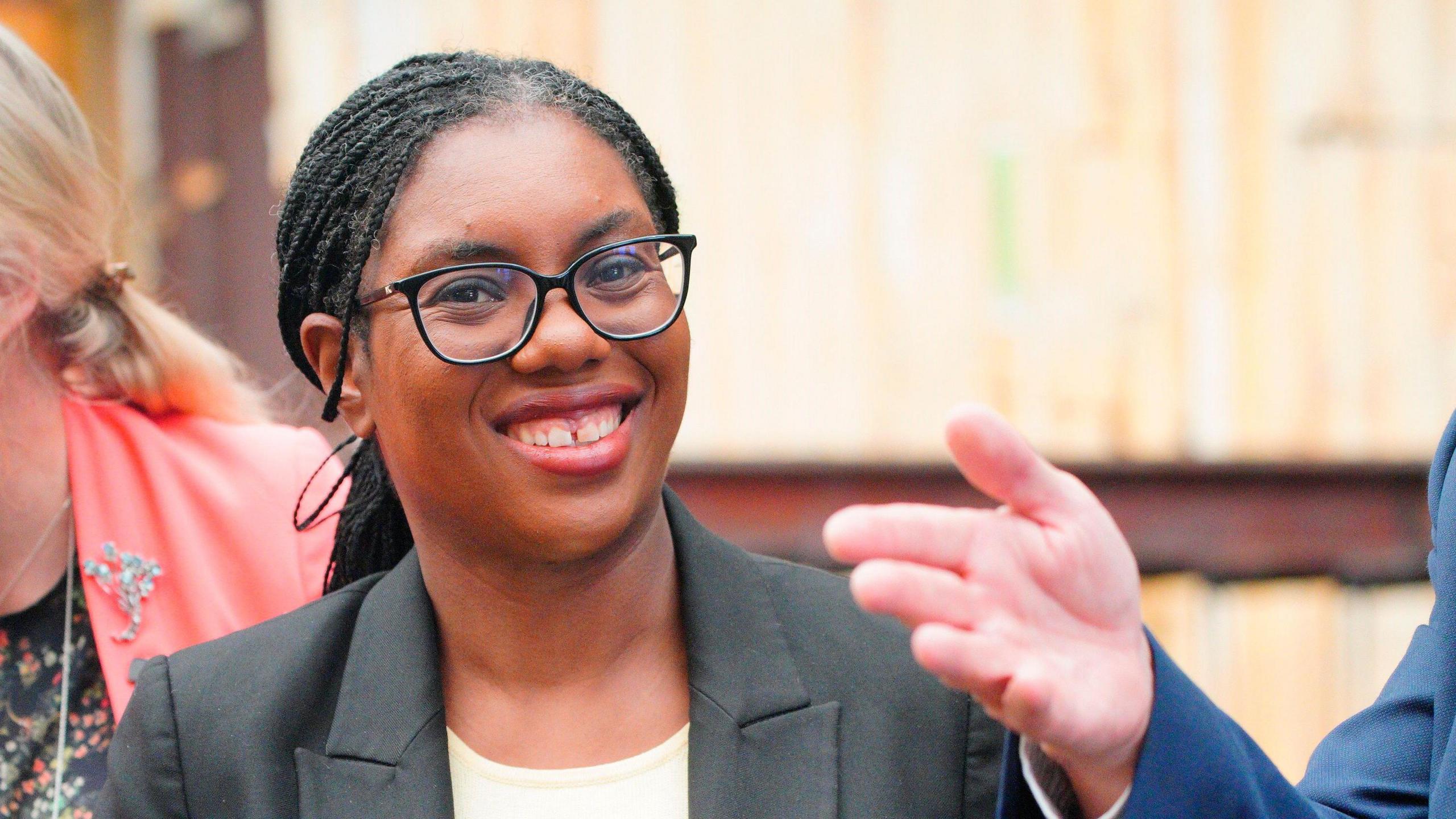 A woman wearing a grey blazer and a white top and smiling at the camera. She is wearing black frame glasses