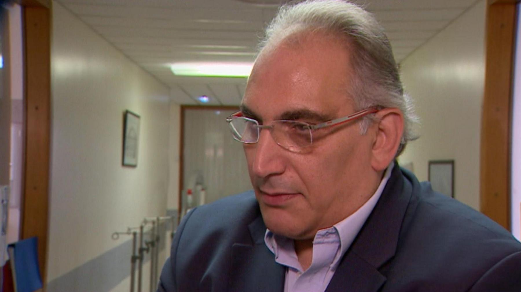 Professor Derek Manas wearing glasses, a dark blue blazer and a purple shirt. He has grey hair, swept back. He is standing in a hospital corridor