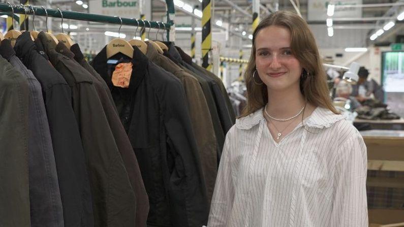 young woman standing in the foreground beside a clothing rail of dark waxed jackets on wooden Barbour hangers. The person is wearing a light striped shirt, hoop earrings and a necklace, and is facing the camera. The setting is a factory or workshop, with industrial lighting, metal framework and workstations visible in the background. Barbour signs can be seen above the clothing rail, and one jacket has an orange tag attached to it. No bags, bins or floor markers are clearly visible.