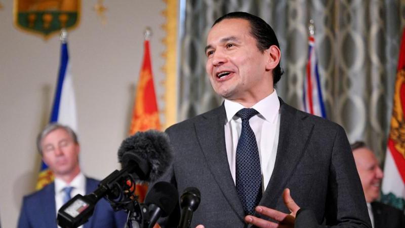 An image showing Wab Kinew speaking in front of several microphones at a press conference with a few of Canada's other premiers behind him. He has black hair that is tied up in a bun, and is wearing a dark charcoal grey suit and tie.