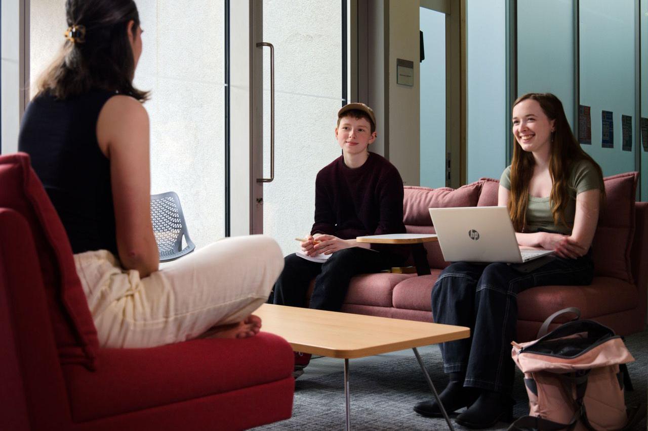 Dr Cole sits with two students for a supervision at the English faculty in Cambridge. They are in a modern indoor meeting area with sofas and a low table. One person sits in the foreground facing away from the camera, while two others sit opposite on a pink sofa, one using a laptop and the other holding a notebook. The setting features glass walls, soft furnishings, and a casual, collaborative atmosphere.