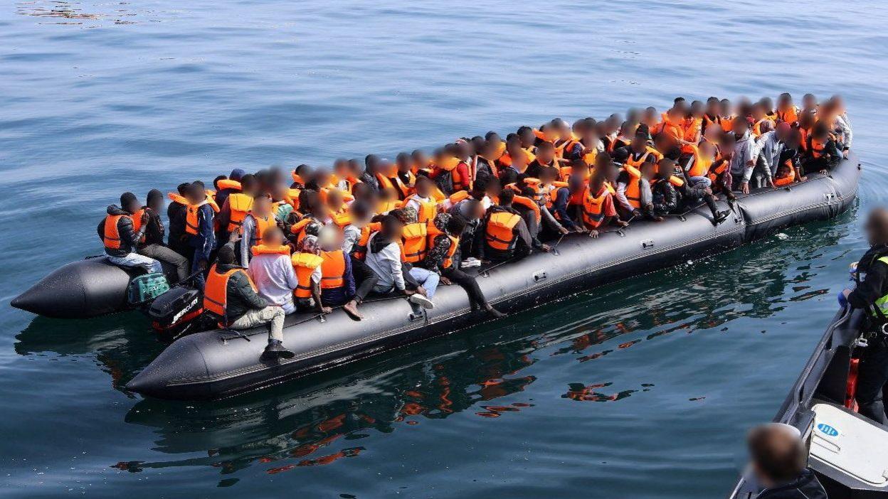 A large number of people in orange lifejackets on an inflatable boat.