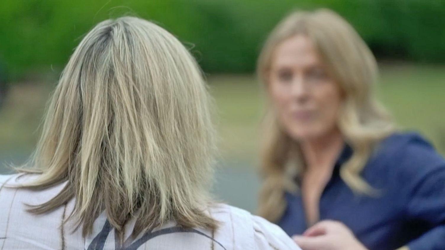 Rear view of Jo with shoulder-length light-coloured hair - she is wearing a light-coloured patterned top. The background is softly blurred. She can be seen facing reporter Sharon Barbour.