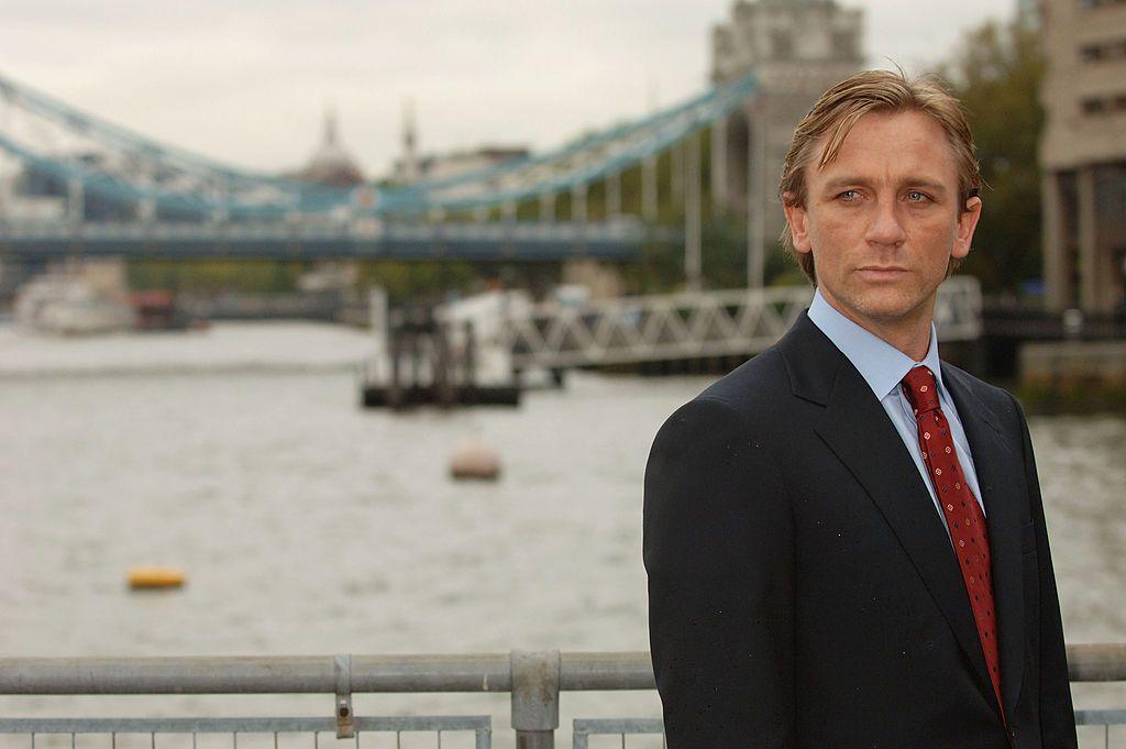 On the right of the frame, Daniel Craig stands wearing a suit on a bridge with the London skyline behind him