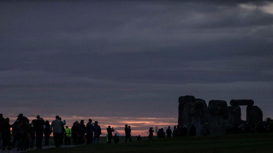 The stones of Stonehenge are visible as black shadows to the right, in front of a dark grey sky with fragments of an orange and pink sunrise visbile towards the horizon. People stand around to the left, some with cameras held up to capture the moment.