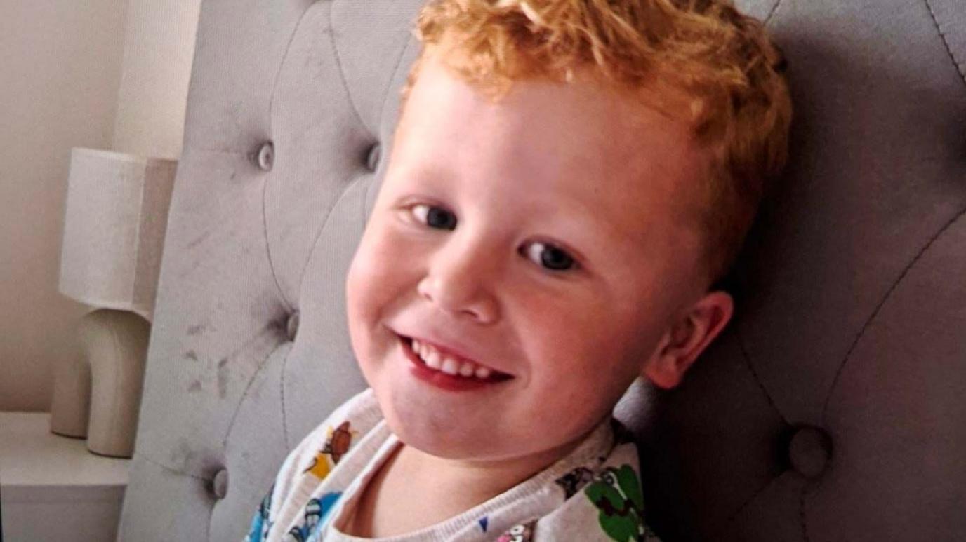 Noah, a three-year-old boy who has ginger hair. He is pictured sitting on a bed with a grey headboard and is looking directly at the camera and smiling.