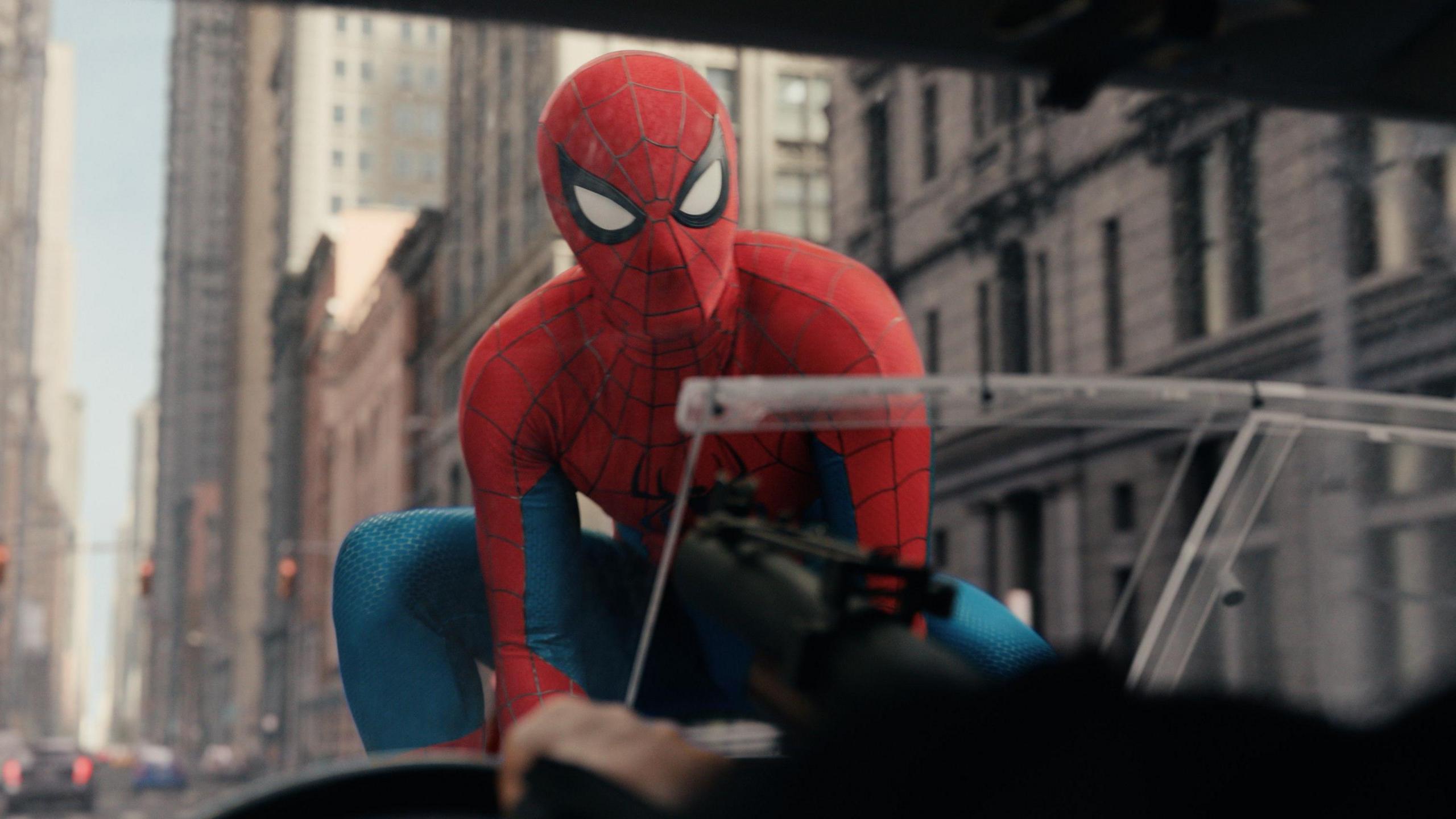Spiderman on the bonnet of a car, with a number of tall stone buildings behind him.
