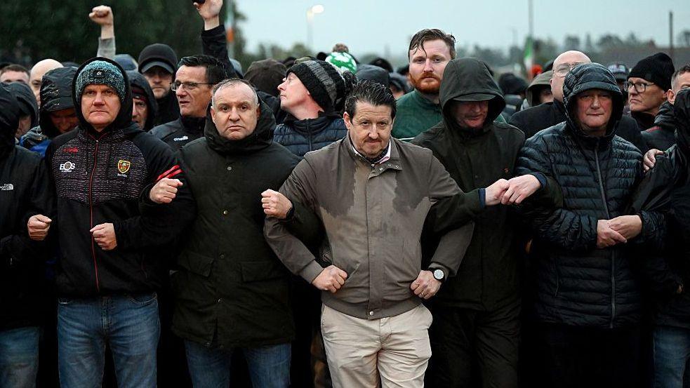 Line of protesters, mainly men, stand in rows and link arms. It's raining and most are wearing coats, some with their hoods up