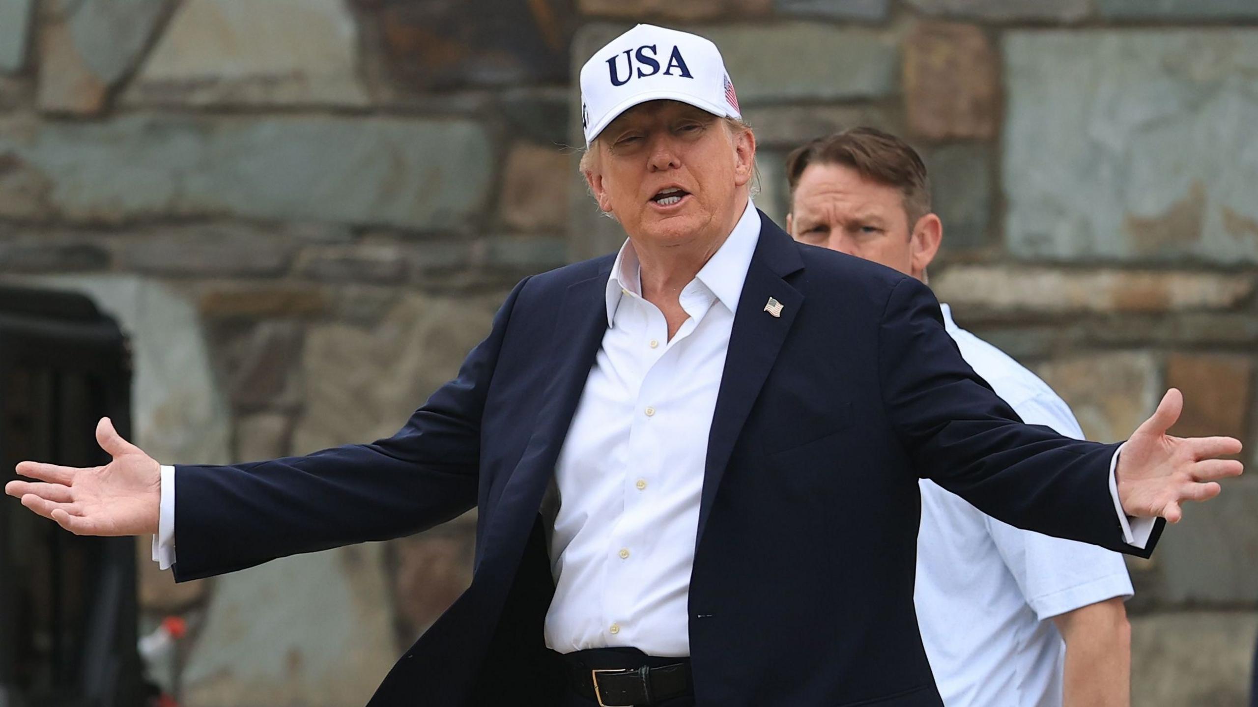 U.S. President Donald Trump attends the Amgen Irish Open at Trump International Golf Links in Doonbeg, Ireland. He is wearing a white USA baseball cap, navy blazer with a white shirt. He has his arms outstretched.