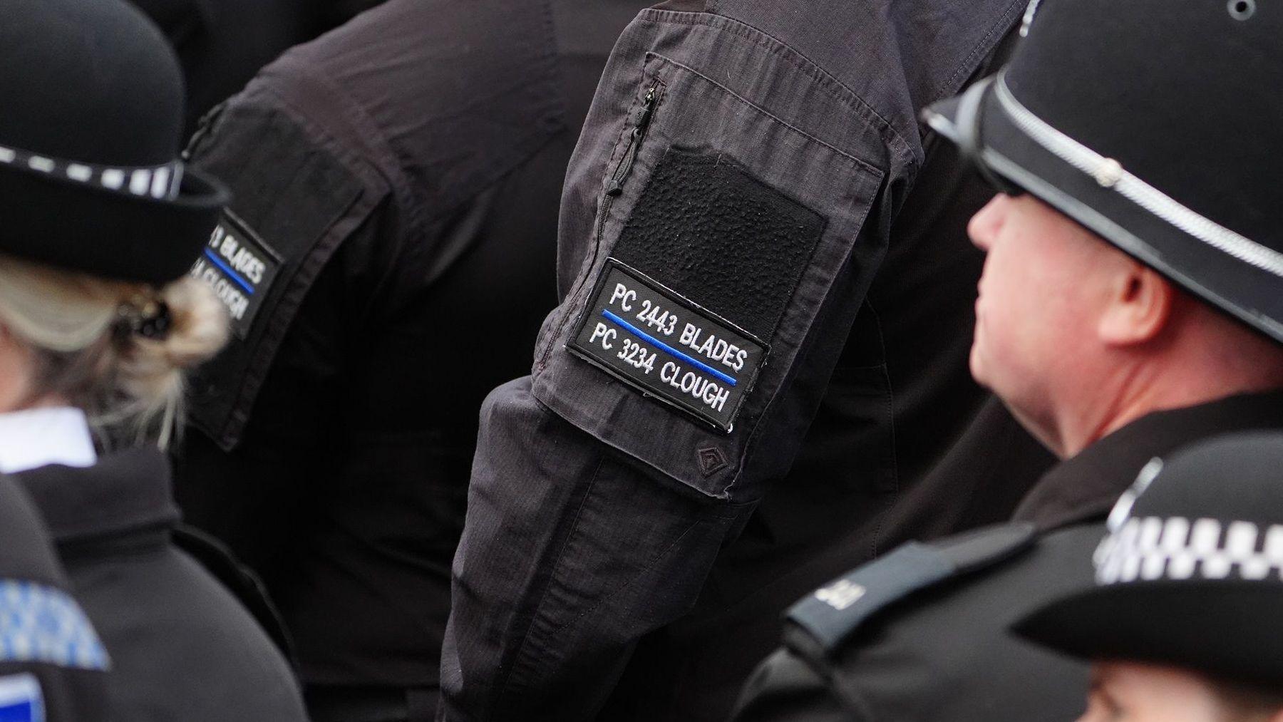 A close-up of the arm of one of the police officers who is in all black and wearing an arm patch which reads PC 2443 Blades and PC 3234 with a blue line through the middle. A police officer in front of them is wearing the same patch which is partially cut off by the head of a female police officer with a round hat on and her blonde hair in a bun.