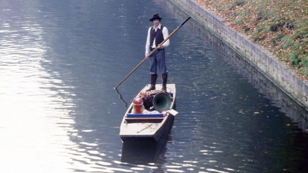 Tom Baker is wearing a black hat, white shirt and black waistcoat, navy trousers and black boots. He is standing on a wooden punt which is on the River Cam. On the right is a wall leading to a field.