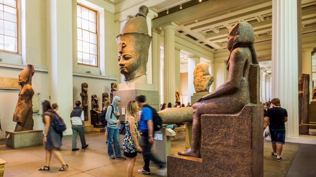 A large room with ornate white pillars. Several people looking at large Egyptian sculptures
