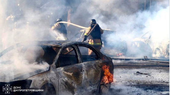Two firefighters direct a hose at billowing smoke in front of them. A burned-out car is on fire in the foreground.