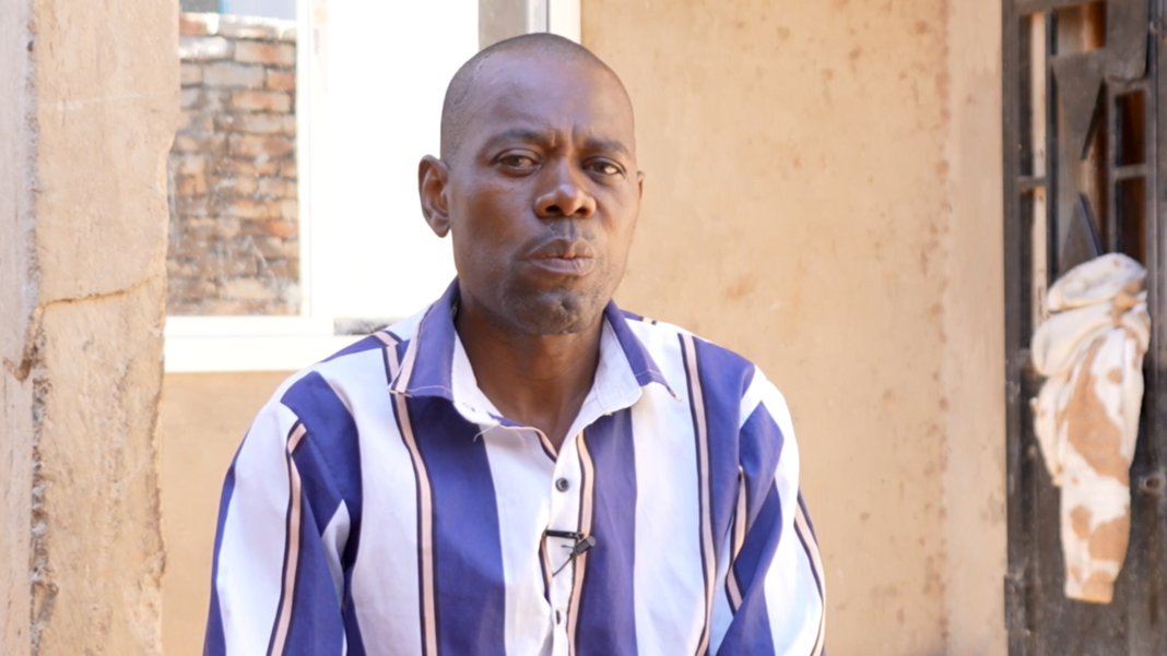 How killings sparked by accusations of ‘genital theft’ are being fuelled by social media A medium shot of an adult man with short hair seated outdoors against a beige plastered wall looking directly into the camera with a serious facial expression. He is wearing a long-sleeved, collared button-down shirt featuring vertical blue, white, and thin pink stripes.