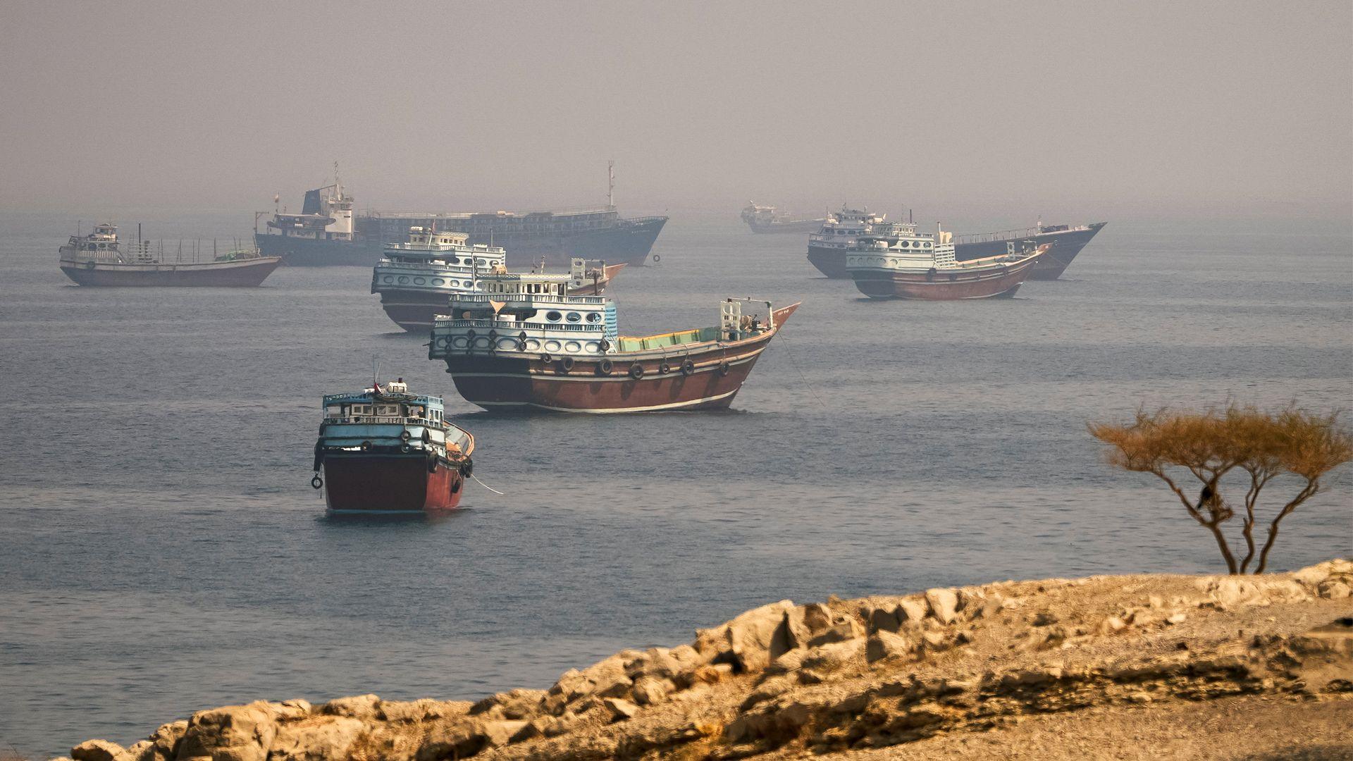 Several ships sit in a large body of water, with fog on the horizon and rocks in the foreground.