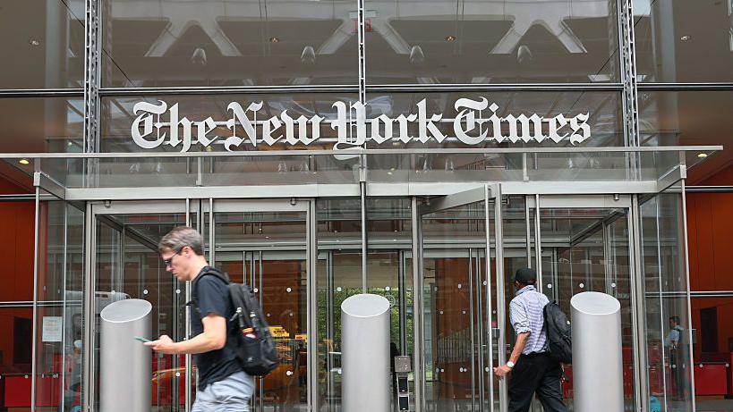 The New York Times building is seen on September 16, 2025 in New York City. The images features the logo of the newspaper, displayed atop the building's front doors, with people walking by.