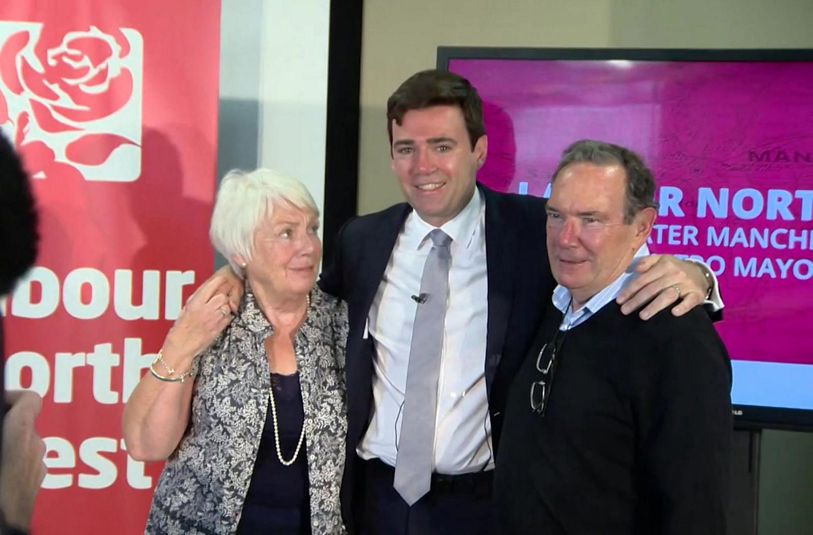 Andy Burnham hugs his parents Eileen and Roy in 2016, when he was selected as the Labour candidate for the Greater Manchester mayoral race. Head and shoulders shot of Burnham, who is smiling in between his mother and father, with his arms draped over their shoulders. He is wearing a black suit, white shirt and grey tie. His mother stands on the left with white hair and a black and white paisley shirt over a black top. His father stands o the right wearing a black jumper over a blue and white open-necked shirt.