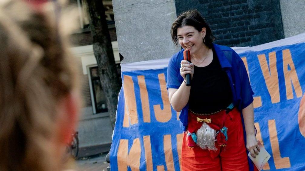 A person with brown hair, holding a mic smiling. She wears red trousers and a blue shirt. She also has a merkin - or pubic hair wig - over her clothes. Behind her is a large blue and orange cloth sign used on a march to celebrate a new Dutch word for labia.