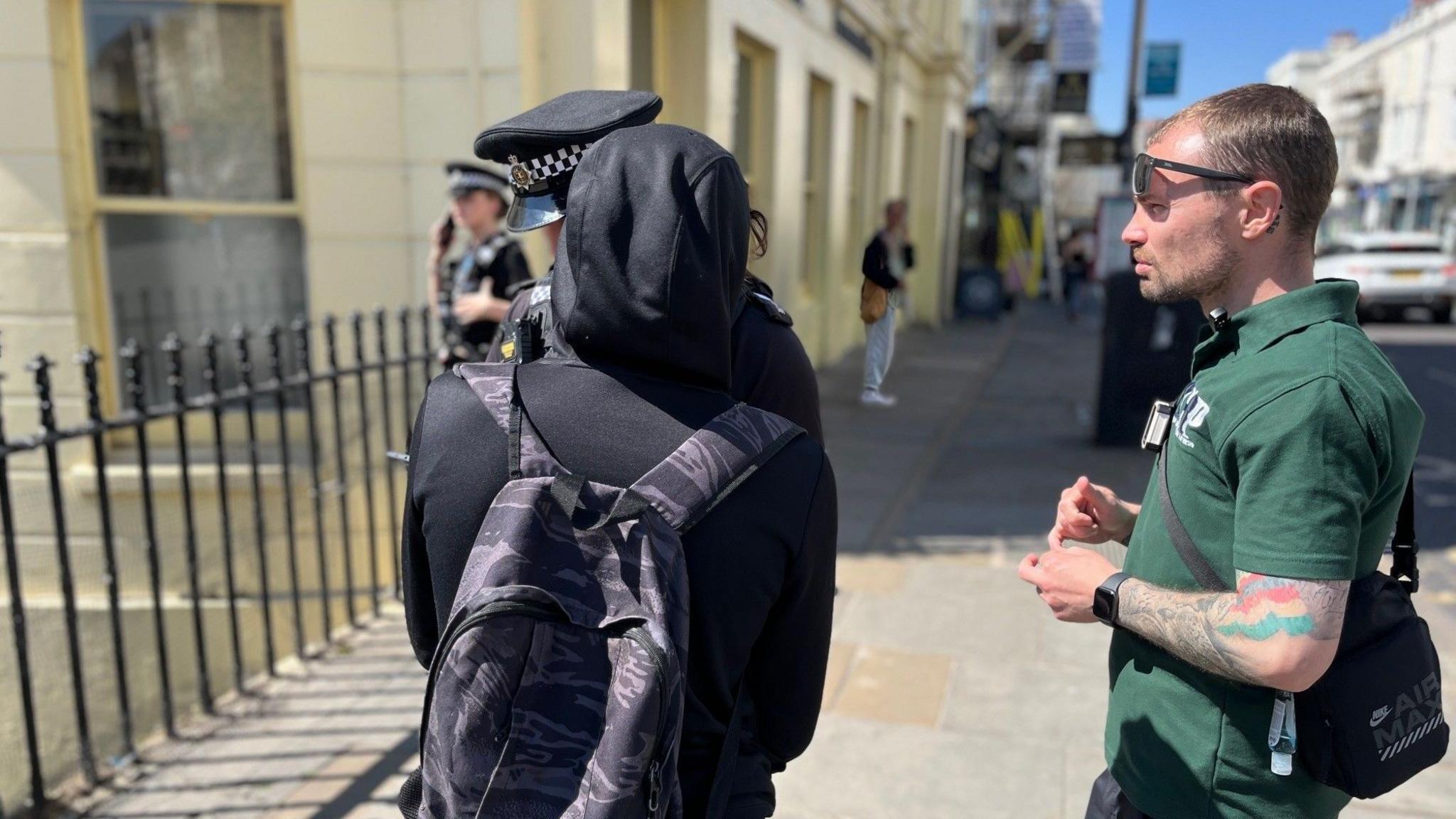 A man with a dark green polo shirt, and tattoos on his arm, is talking to a police officer and a person in a black hoodie with their back to the camera.